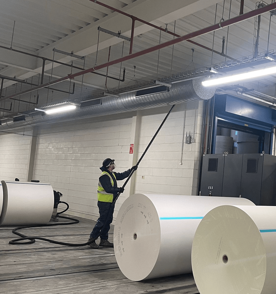 APC technician using long-reach vacuum to clean ceiling pipes near large paper rolls